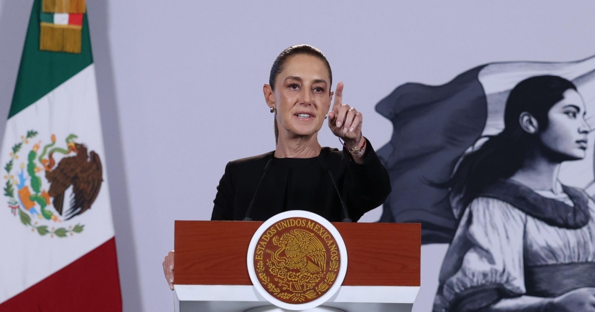 presidenta de México, Claudia Sheinbaum Pardo, de pie detrás del podio oficial de las conferencias mañaneras en Palacio Nacional. Vestida con un saco oscuro, levanta el dedo índice de su mano izquierda mientras habla. A su derecha (izquierda de la imagen) se aprecia la bandera nacional mexicana y a la derecha una ilustración de fondo en escala de grises con la imagen institucional de heroínas de la patria. El podio luce el escudo de los Estados Unidos Mexicanos sobre acabado de madera.