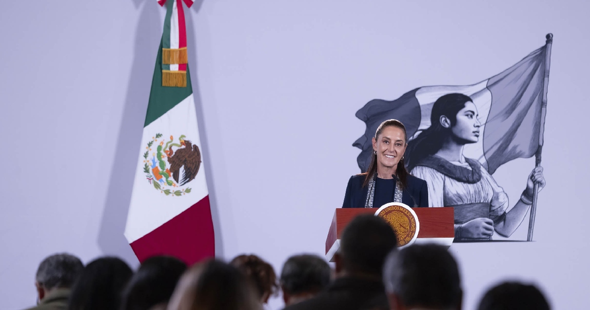 presidenta de México, Claudia Sheinbaum Pardo, al frente del pódium en el Salón Tesorería de Palacio Nacional durante su conferencia de prensa matutina («La Mañanera del Pueblo»). A su derecha se aprecia la Bandera Nacional de México, y en el fondo destaca la ilustración gráfica de la joven heroína mexicana de la patria. En primer plano, de espaldas y en silueta, se observan los periodistas y reporteros asistentes al encuentro informativo.