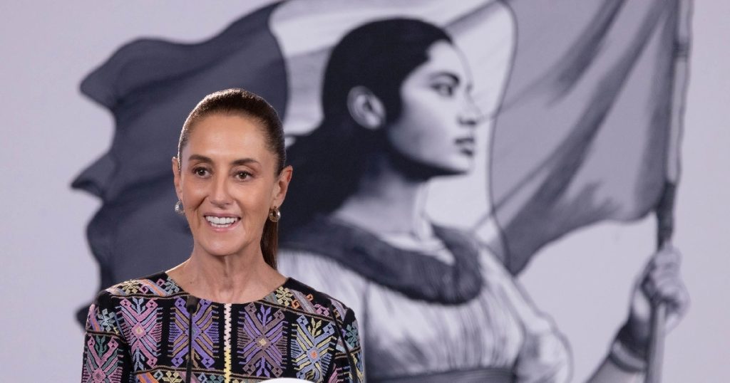 Primer plano de la presidenta de México, Claudia Sheinbaum Pardo, sonriendo y expresándose durante su conferencia de prensa matutina «La Mañanera del Pueblo» en Palacio Nacional. Viste un blusón bordado tradicional con motivos artesanales geométricos en tonos morados, rosas y amarillos. Al fondo, en segundo plano, destaca el emblema institucional con la ilustración en tonos grises de la joven heroína mexicana sosteniendo la bandera.