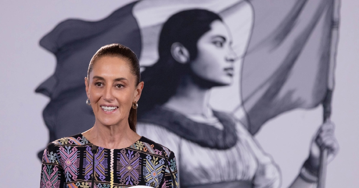 Primer plano de la presidenta de México, Claudia Sheinbaum Pardo, sonriendo y expresándose durante su conferencia de prensa matutina «La Mañanera del Pueblo» en Palacio Nacional. Viste un blusón bordado tradicional con motivos artesanales geométricos en tonos morados, rosas y amarillos. Al fondo, en segundo plano, destaca el emblema institucional con la ilustración en tonos grises de la joven heroína mexicana sosteniendo la bandera.