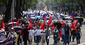 una multitud marchando sobre una avenida principal ( Paseo de la Reforma / Juárez en la Ciudad de México). El contingente de manifestantes porta numerosas banderas rojas, moradas y pancartas, mientras caminan hacia adelante portando sombrillas para protegerse del sol.