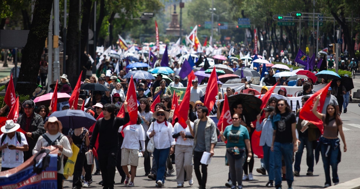 una multitud marchando sobre una avenida principal ( Paseo de la Reforma / Juárez en la Ciudad de México). El contingente de manifestantes porta numerosas banderas rojas, moradas y pancartas, mientras caminan hacia adelante portando sombrillas para protegerse del sol.