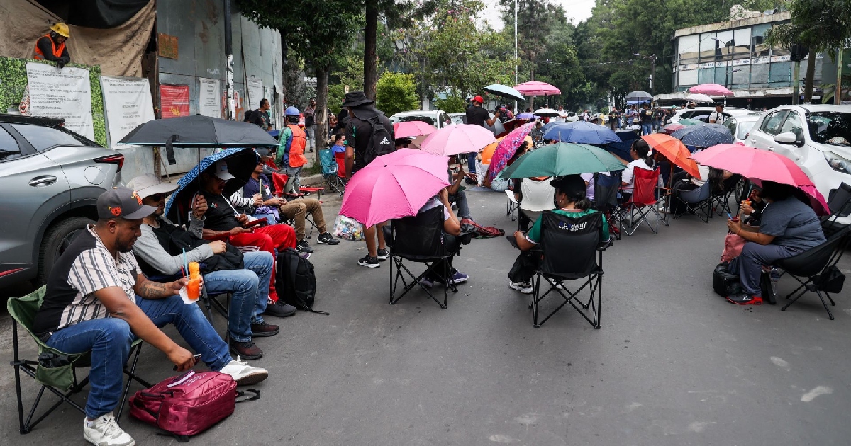 un bloqueo vial y manifestación pacífica sobre una calle de la Ciudad de México. Decenas de personas se encuentran sentadas en sillas plegables de camping a lo largo del asfalto, cubriéndose del sol con sombrillas de diversos colores