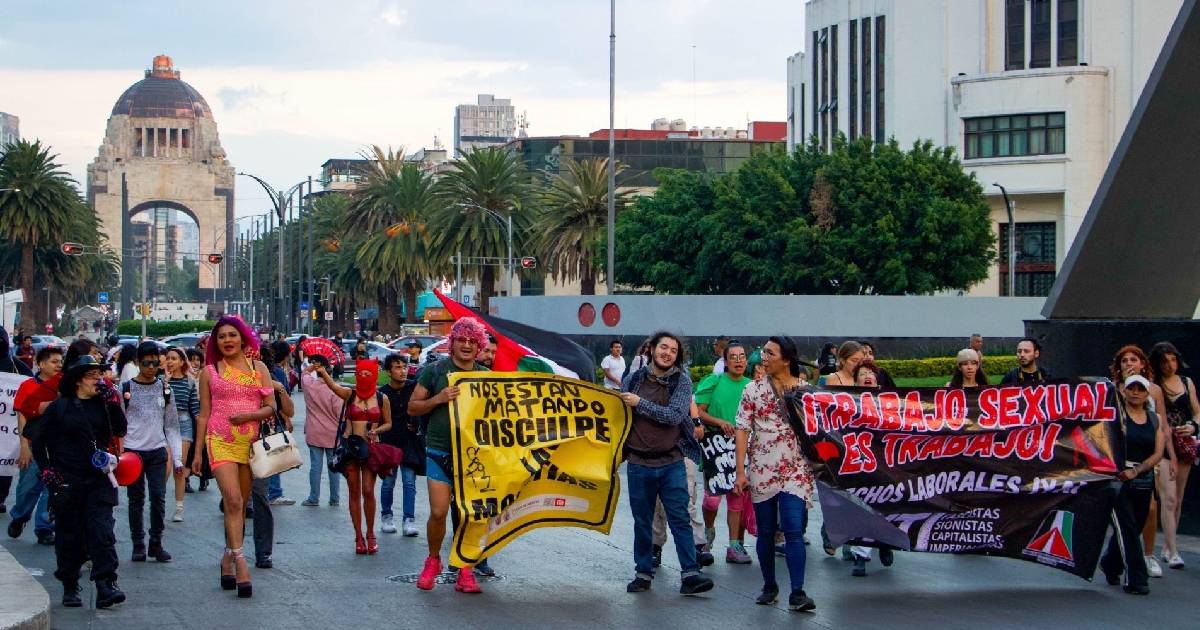 Plano general de una manifestación y marcha de contingentes de trabajadoras sexuales y colectivos LGBTTTIQA+ avanzando sobre una avenida en la Ciudad de México. En la vanguardia del contingente se observan activistas desplegando lona y mantas con consignas como «¡Trabajo sexual es trabajo!» y «Nos están matando, disculpe las molestias». Al fondo de la avenida destaca el Monumento a la Revolución bajo un cielo nublado, flanqueado por palmeras y edificios del Centro Histórico.