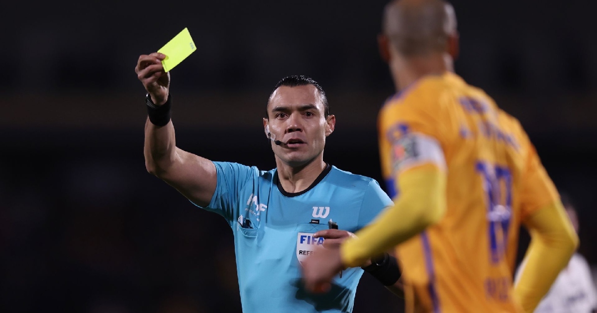 árbitro profesional de fútbol Marco Antonio Ortiz Nava vistiendo el uniforme oficial azul turquesa de la Liga MX con el parche FIFA REFEREE. El silbante sostiene en alto una tarjeta amarilla con la mano derecha mientras mira fijamente hacia un futbolista de espaldas vestida con camiseta amarilla y azul (Tigres UANL).