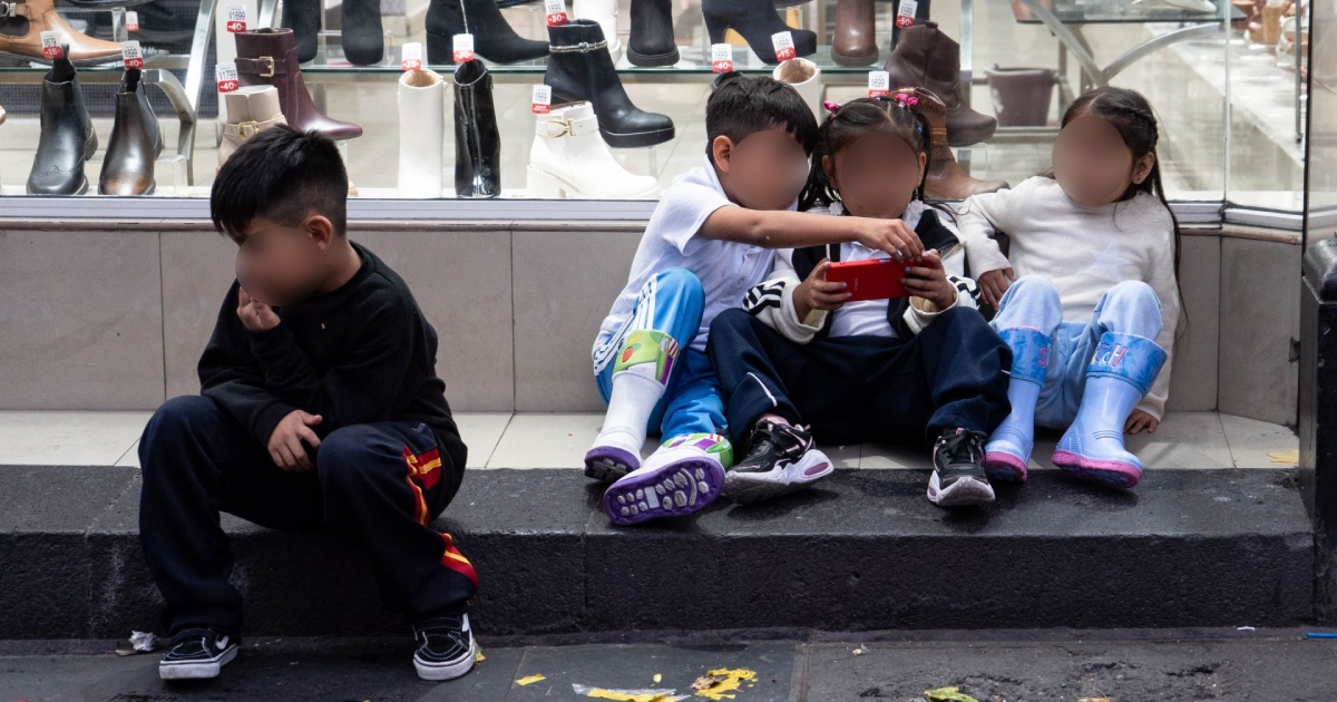 cuatro niñas y niños sentados sobre el escalón del mostrador de una tienda de calzado en la vía pública. A la izquierda, un niño en pants oscuro permanece sentado mirando hacia el suelo.