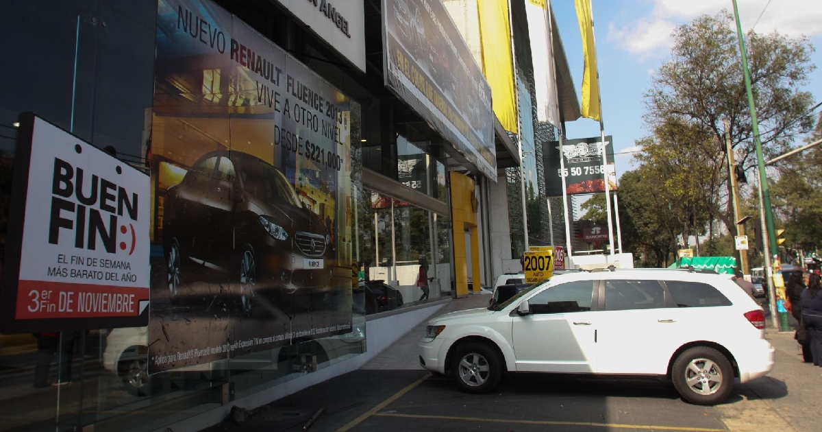 exterior de una agencia concesionaria de automóviles en la Ciudad de México. En la fachada de cristal destaca un gran cartel publicitario con la promoción del «El Buen Fin» («El fin de semana más barato del año») junto a un anuncio de un automóvil Renault Fluence. En la explanada frontal y sobre la banqueta se encuentra estacionada una camioneta tipo SUV de color blanco, mientras al fondo se aprecian letreros de financiamiento, semáforos y transeúntes sobre la avenida.