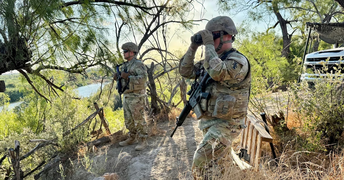 dos soldados estadounidenses de la Guardia Nacional vistiendo uniformes de camuflaje, cascos, chalecos tácticos y portando rifles de asalto en un puesto de observación elevado. El soldado en primer plano mira a través de unos binoculares hacia la cuenca de un río (Río Bravo/Río Grande) rodeado de vegetación y maleza silvestre.