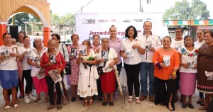 Fotografía grupal de plano general al aire libre en un soportal o plaza comunitaria que muestra a un numeroso grupo de mujeres indígenas y pobladoras locales posando sonrientes junto a funcionarios estatales de Puebla.