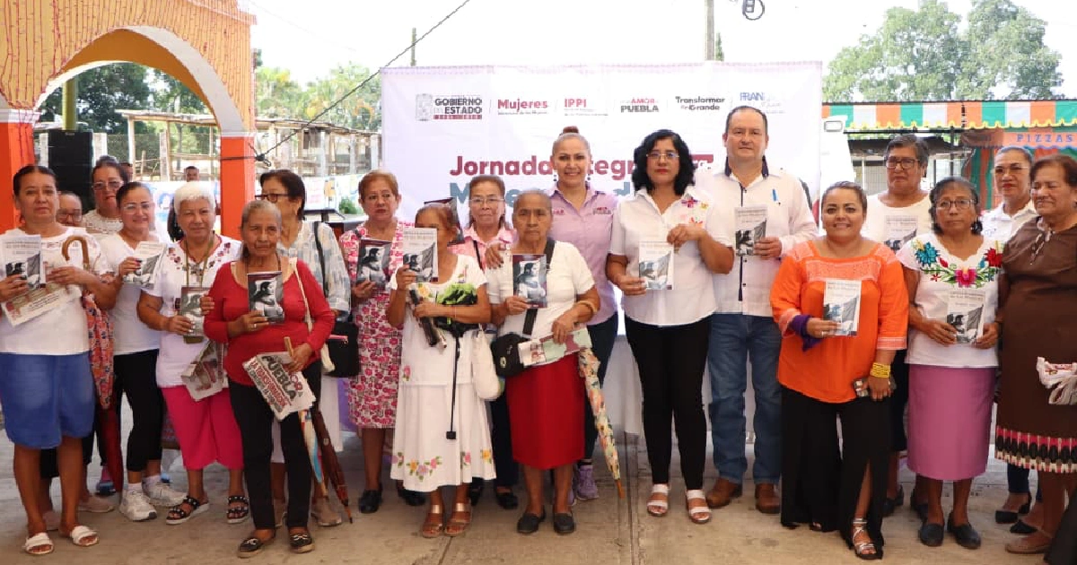 Fotografía grupal de plano general al aire libre en un soportal o plaza comunitaria que muestra a un numeroso grupo de mujeres indígenas y pobladoras locales posando sonrientes junto a funcionarios estatales de Puebla.