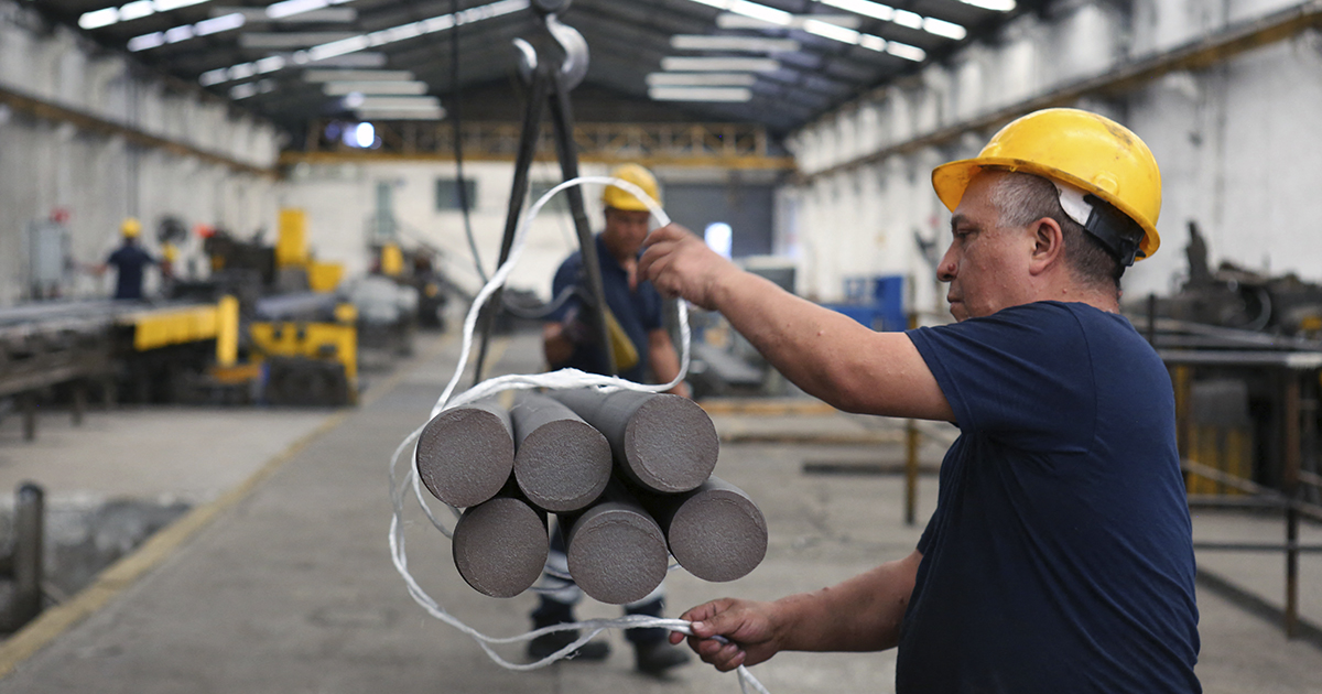 Plano medio en una nave industrial metalúrgica o de procesamiento minero. Un operario con casco de seguridad amarillo y camiseta azul maneja un conjunto de barras metálicas cilíndricas de gran grosor (aleaciones de niobio/acero especial) sujetas por una eslinga y suspendidas de un gancho de grúa.