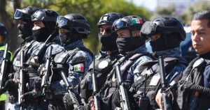 uerzas de operaciones especiales y elementos policiales fuertemente armados. Visten uniformes tácticos con camuflaje pixelado gris y azul, chalecos antibalas, cascos con antiparras y pasamontañas negros que cubren la mitad inferior de sus rostros. Llevan parches con la bandera de México en los brazos y cascos, sosteniendo sus armas largas de cargo en posición de firmeza durante un despliegue operativo.