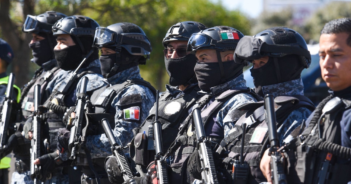 uerzas de operaciones especiales y elementos policiales fuertemente armados. Visten uniformes tácticos con camuflaje pixelado gris y azul, chalecos antibalas, cascos con antiparras y pasamontañas negros que cubren la mitad inferior de sus rostros. Llevan parches con la bandera de México en los brazos y cascos, sosteniendo sus armas largas de cargo en posición de firmeza durante un despliegue operativo.