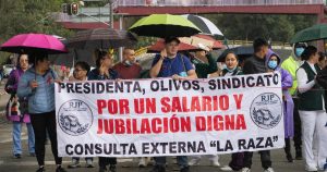manifestación de trabajadores de la salud del Instituto Mexicano del Seguro Social (IMSS) marchando bajo la lluvia en una avenida vial. En primer plano, los manifestantes sostienen una manta blanca con letras negras y rojas donde se lee: «PRESIDENTA, OLIVOS, SINDICATO POR UN SALARIO Y JUBILACIÓN DIGNA CONSULTA EXTERNA “LA RAZA”», acompañada de los logotipos de la organización RJP (Recuperemos el Régimen de Jubilaciones y Pensiones).