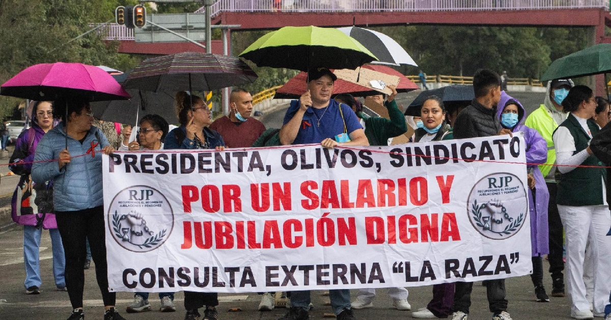 manifestación de trabajadores de la salud del Instituto Mexicano del Seguro Social (IMSS) marchando bajo la lluvia en una avenida vial. En primer plano, los manifestantes sostienen una manta blanca con letras negras y rojas donde se lee: «PRESIDENTA, OLIVOS, SINDICATO POR UN SALARIO Y JUBILACIÓN DIGNA CONSULTA EXTERNA “LA RAZA”», acompañada de los logotipos de la organización RJP (Recuperemos el Régimen de Jubilaciones y Pensiones).