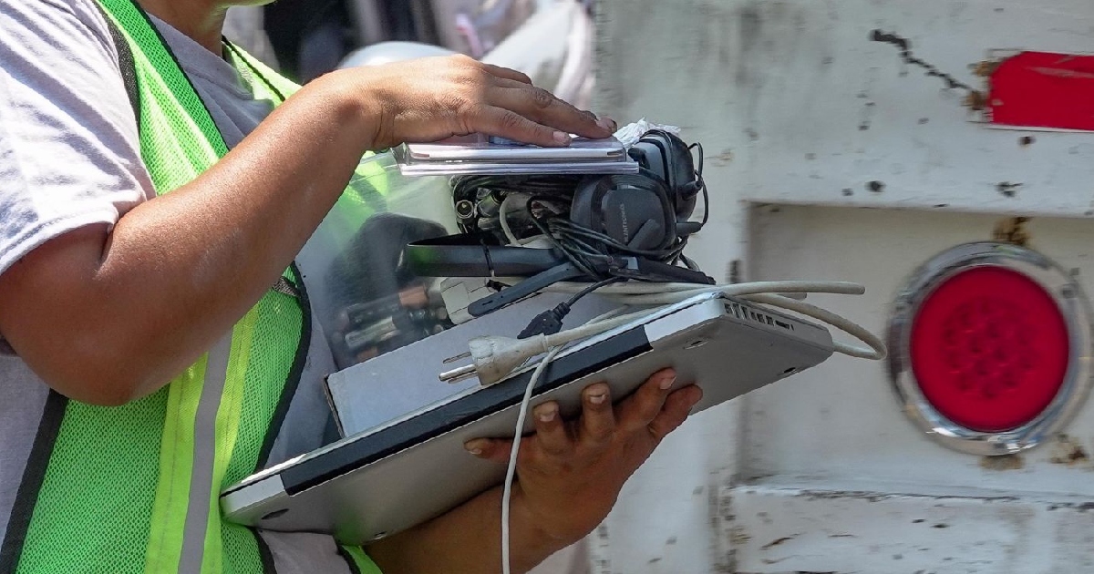 un trabajador que viste un chaleco de seguridad verde reflejante cargando un contenedor transparente y varios dispositivos electrónicos obsoletos. Entre los artículos se aprecian una computadora portátil (laptop) gris plateada con sus cables de alimentación, audífonos con micrófono, pilas alcalinas en un contenedor plástico y periféricos diversos listos para ser ingresados a un camión de recolección de residuos.