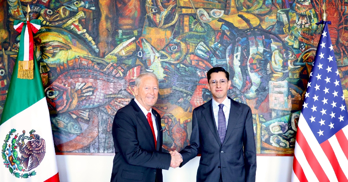 saludo de manos (estrechamiento de manos) entre el diplomático estadounidense Ronald Johnson y Roberto Velasco Álvarez, jefe de la Unidad para América del Norte de la Secretaría de Relaciones Exteriores (SRE). Ronald Johnson viste un traje oscuro con corbata roja, mientras que Roberto Velasco luce un traje gris oscuro, camisa blanca y corbata azul estampada. Ambos posan sonrientes de frente a la cámara. Flanqueando la escena se encuentran la Bandera Nacional de México (a la izquierda) y la Bandera de los Estados Unidos (a la derecha). Al fondo destaca una obra pictórica o mural con motivos marinos y pesqueros en tonalidades cálidas y coloridas.
