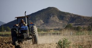 A la izquierda un hombre un tractor color azul circulando sobre un terreno plano y árido, al fondo se observa una montaña algo seca.