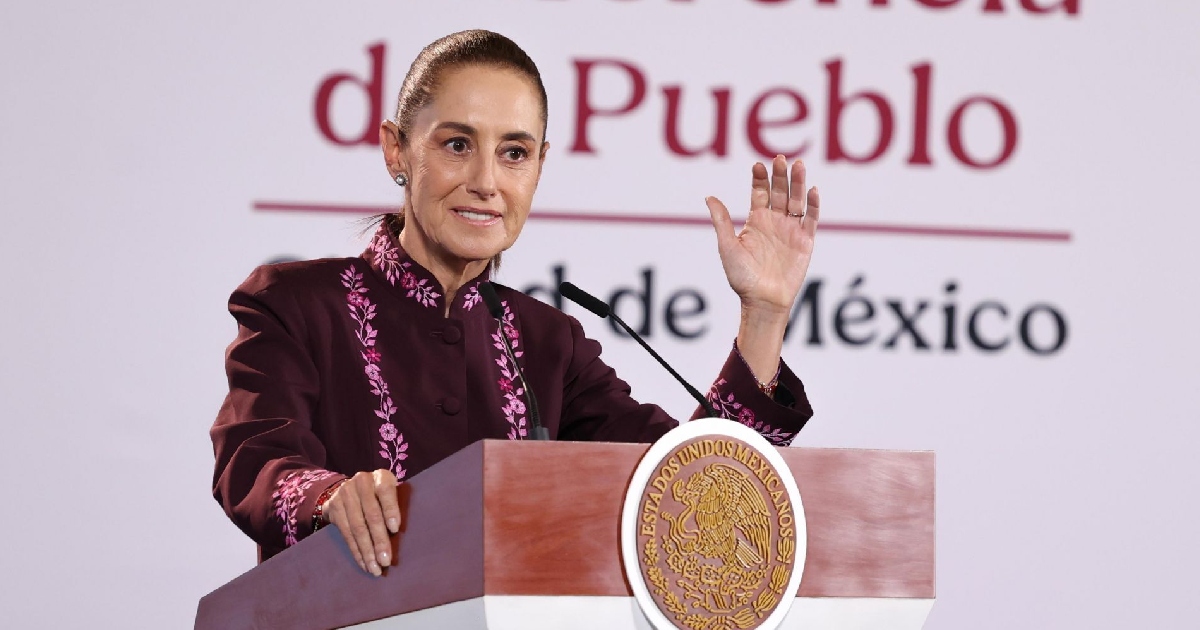 Plano medio horizontal de la presidenta Claudia Sheinbaum Pardo durante una intervención en la conferencia matutina «La Mañanera del Pueblo». Viste un saco/chaqueta de tono guinda o vino tinto con bordados artesanales florales en tono rosa a lo largo de la solapa y los puños. Se encuentra posicionada detrás del presídium de madera con el Escudo Nacional en dorado y sostiene la mano izquierda levantada con un gesto explicativo hacia la audiencia. Al fondo destaca la mampara oficial blanca con la tipografía institucional en guinda.