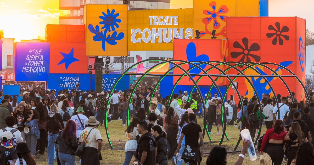 Plano general en perspectiva durante el atardecer del pórtico de entrada y explanada del festival Tecate Comuna. La imponente estructura de acceso está compuesta por paneles tridimensionales de vivos colores amarillo, azul y naranja con tipografía de estilo psicodélico donde se lee «TECATE COMUNA» y frases decorativas como «QUE SE SIENTA EL POWER MEXICANO».