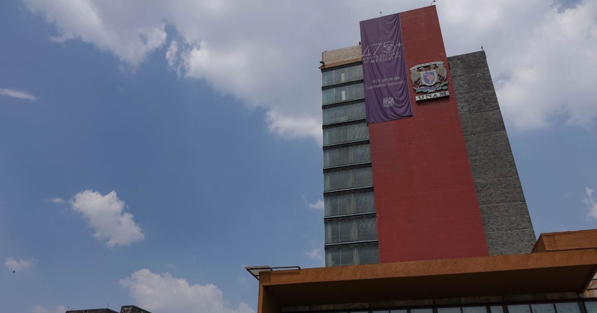 Vista arquitectónica en contrapicado de la Torre de Rectoría de la Universidad Nacional Autónoma de México (UNAM) en Ciudad Universitaria. En la fachada roja del emblemático edificio luce el escudo oficial de la UNAM junto a un pendón morado conmemorativo con la leyenda «475+ Universidad de México, 475 años de concebir futuros». El edificio destaca sobre un cielo azul despejado con nubes blancas dispersas.